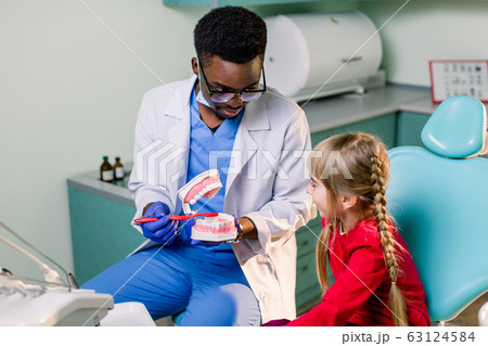 Happy smiling black dentist tells Caucasian little girl how to brush her teeth. Caries prevention, Dentistry, teeth hygiene concept 63124584