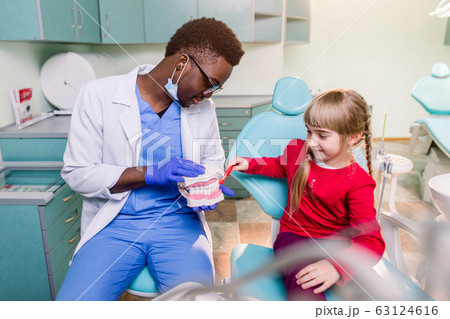 The child girl in dentist's chair having fun while brushing teeth with a male African American doctor 63124616