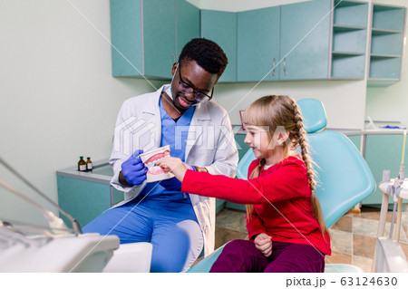 A smiling young African American dentist holds artificial jaws in his arms, and a little girl brushes with toothbrush teeth layout. Pediatric dentistry concept 63124630