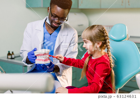 A smiling young African American dentist holds artificial jaws in his arms, and a little girl brushes with toothbrush teeth layout. Pediatric dentistry concept 63124631
