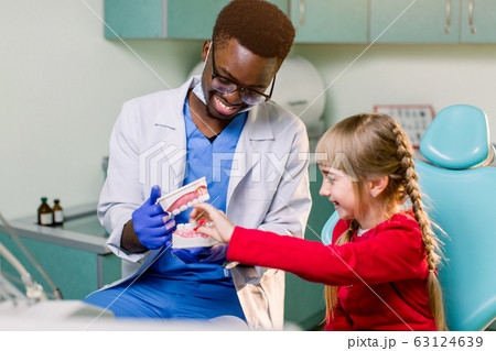 African dentist shows how to brush teeth to a small Caucasian girl a patient, using a artificial jaws. Dentistry, teeth hygiene concept 63124639