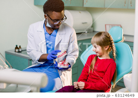 Little patient at the dentist's office. Young African American dentist talking with patient small girl holding artificial jaws in his hands. 63124649