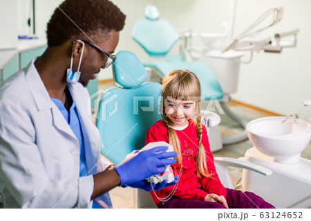 Little patient at the dentist's office. Young African American dentist talking with patient small girl holding artificial jaws in his hands. 63124652