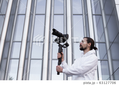 Young Professional videographer holding professional camera on 3-axis gimbal stabilizer. Pro equipment helps to make high quality video without shaking. Cameraman wearing white hoodie making a videos. 63127706