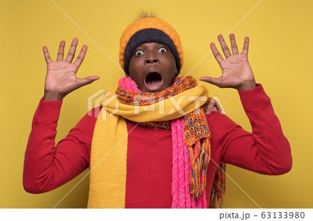 Scared african american man in several hats and scarfs on yellow background. Scared african american man in several hats and scarfs on yellow background. 63133980
