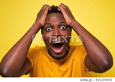 Amazed African American man in yellow clothes holding hands in surprised gesture Amazed African American man in yellow clothes holding hands in surprised gesture 63134043