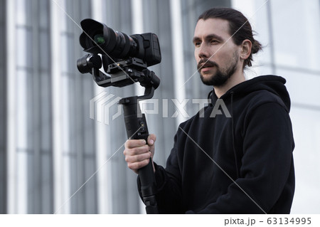 Young Professional videographer holding professional camera on 3-axis gimbal stabilizer. Pro equipment helps to make high quality video without shaking. Cameraman wearing white hoodie making a videos. 63134995