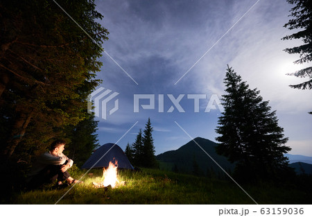 Guy is sitting by the fire on background of tent and spruce forest enjoying magical view mountains in the evening. 63159036