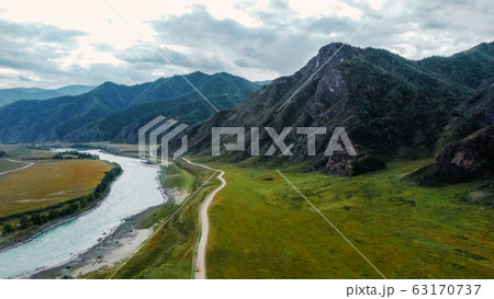 The road next to the river along the mountain ranges. Mountain landscape aerial view from a drone. Altai The road next to the river along the mountain ranges. Mountain landscape aerial view from a drone. Altai 63170737