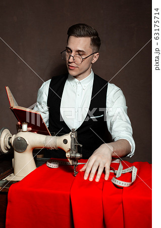 tailor in round glasses sits at a table with a sewing machine 63175714