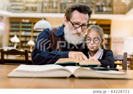 Elderly man grandfather and his granddaughter reading exciting book together while sitting in the library, on the background of vintage book shelves. Education, family reading Elderly man grandfather and his granddaughter reading exciting book together while sitting in the library, on the background of vintage book shelves. Education, family reading 63226721