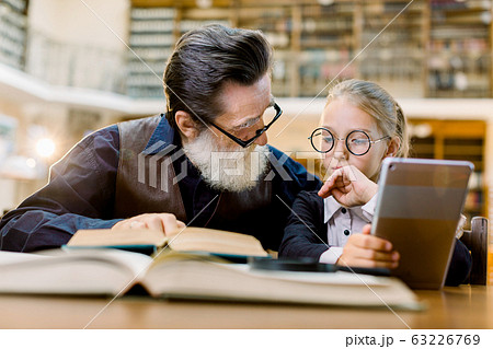 Smart pretty girl in eyeglasses and old man teacher having a lesson and discussion in vintage library. Girl holds digital tablet or ebook reader and man shows some information in book Smart pretty girl in eyeglasses and old man teacher having a lesson and discussion in vintage library. Girl holds digital tablet or ebook reader and man shows some information in book 63226769