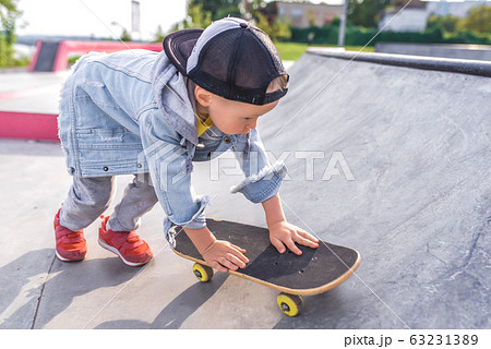 little boy 4-5 years old, rides on skateboard, in summer in city on sports field sits. Casual wear denim and baseball cap. Free space for copy text. The first training lessons. 63231389