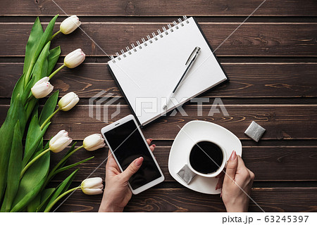 White tulips on a wooden table with an empty notebook, smartphone and a Cup of coffee in women's hands 63245397