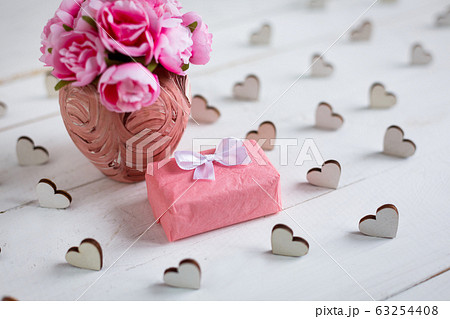 Small gift wrapped in paper with wooden hearts around and pink flowers in vase on white wooden background. 63254408