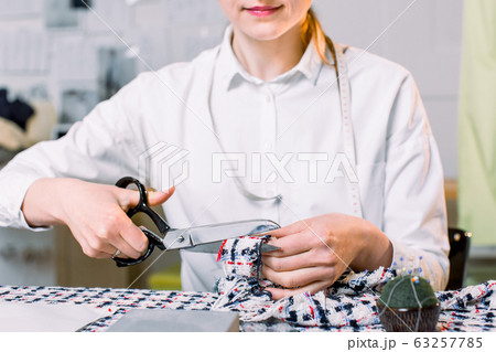 Smiling cute young woman seamstress sitting at the working table and cutting fabric with scissors 63257785