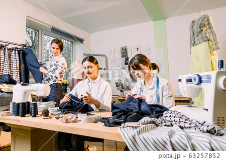 Female fashion designer standing near clothing rack with fashionable stylish handmade clothes. Two young girls dressmakers working on new pants collection for clients in cozy workshop studio 63257852