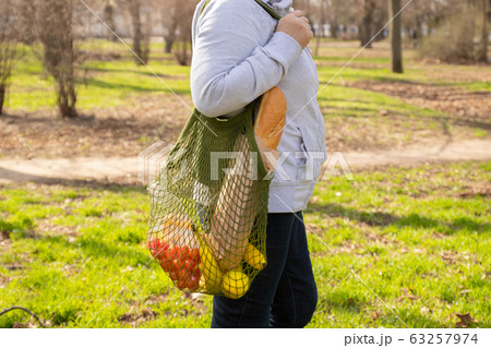 Female hand holds a green ecological cotton mesh bag with a bunch of organic fruits, vegetables and groceries: tomato, bananas, cheese and apples. Zero waste concept. Background, copy space, close up. Female hand holds a green ecological cotton mesh bag with a bunch of organic fruits, vegetables and groceries: tomato, bananas, cheese and apples. Zero waste concept. Background, copy space, close up. 63257974