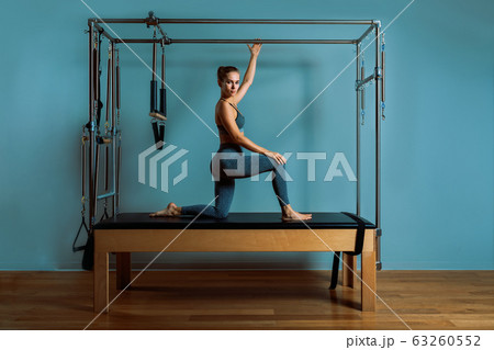 Young girl doing pilates exercises with a reformer bed. Beautiful slim fitness trainer on reformer gray background, low key, art light. Fitness concept Young girl doing pilates exercises with a reformer bed. Beautiful slim fitness trainer on reformer gray background, low key, art light. Fitness concept 63260552