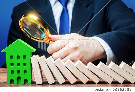 A businessman is studying the consequences of a domino falling on a house. Stress resistance of business and structures. Reliability and quality. Analysis. Real estate insurance and risk management. 63262585