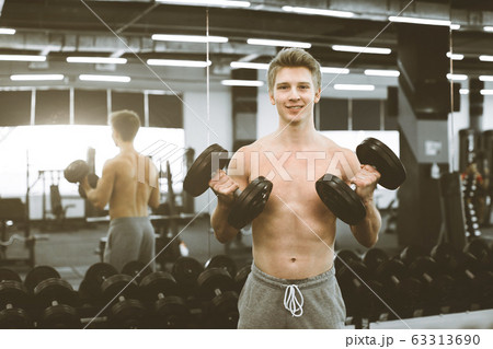 Handsome smiling young blond sportsman is working out with dumbbells, on the background of modern fitness gym 63313690