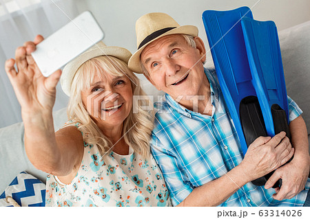 Senior couple sitting in travel agency sitting wearing hats husband holding flippers while wife taking selfie photos on smartphone smiling happy Senior couple sitting in travel agency sitting wearing hats husband holding flippers while wife taking selfie photos on smartphone smiling happy 63314026