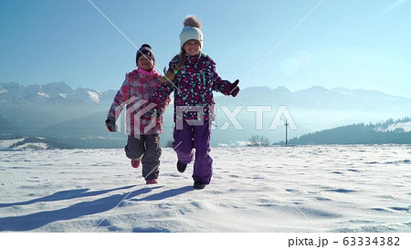 Charming kids in outwear running on snowy terrain with beautiful mountains on background in sunshine Charming kids in outwear running on snowy terrain with beautiful mountains on background in sunshine 63334382
