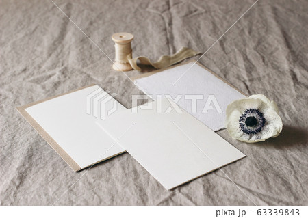 Wedding stationery mock-up scene. Blank greeting cards on linen tablecloth background with white anemone flower and silk ribbon. Feminine still life table composition. High angle view, no people. 63339843