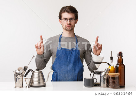 Barista, cafe worker and bartender concept. Portrait of upset grumpy and displeased young male employee feeling unsatisfied and jealous pointing up while standing working bar counter, make coffee Barista, cafe worker and bartender concept. Portrait of upset grumpy and displeased young male employee feeling unsatisfied and jealous pointing up while standing working bar counter, make coffee 63351434