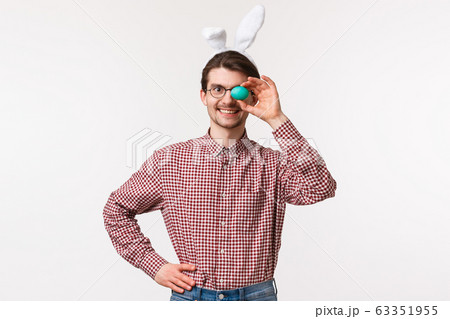 Traditions, religious holidays, celebration concept. Waist-up portrait of cheerful funny caucasian man in rabbit ears, holding one painted egg and smiling satisfied, prepared Easter day meal 63351955