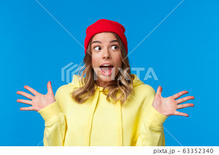 Girl discussing stunning news, latest college gossips, spread hands sideways amused, look away gasping wondered, having interesting conversation, wear red beanie and yellow hoodie 63352340