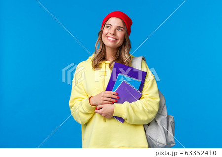 Dreamy cute smart college girl, female student in hipster red beanie, yellow hoodie, looking thoughtful upper left corner imaging summer holidays, smiling holding notebooks and backpack 63352410