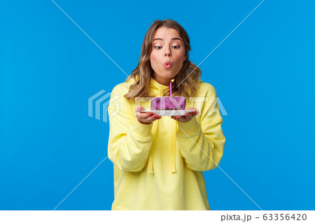 Celebration, party and lifestyle concept. Silly cute european blond woman in yellow hoodie, making wish holding fruit cake blowing out b-day candle, standing blue background at birthday party 63356420