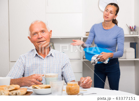 Mature father drinking tea in the kitchen. Adult daughter does cleaning in the apartment 63377151