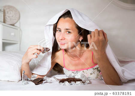 Adult woman secretly eating chocolate under sheets in bed 63378191