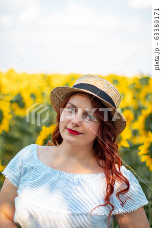 Young red hair Caucasian girl in summer cotton dress and straw hat in a field of yellow sunflowers on a sunny day. 63389171