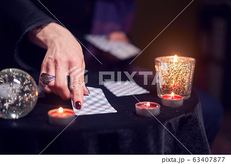 Close-up of female fortuneteller's hands with fortune-telling cards at table with candles Close-up of female fortuneteller's hands with fortune-telling cards at table with candles 63407877