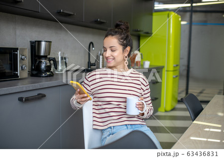 Young woman sitting in the kitchen, drinking coffee, checking phone 63436831