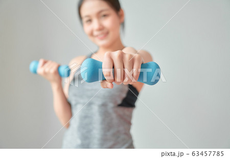 Smiley Asian woman wearing sportswear pumping up muscles with blue dumbbell in living room. Smiley Asian woman wearing sportswear pumping up muscles with blue dumbbell in living room. 63457785