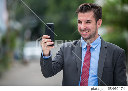 Young happy Hispanic businessman smiling while using phone outdoors 63460474