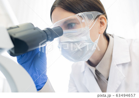 Female researcher in gloves, mask and protective eyewear bending over microscope 63494657