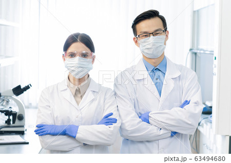 Two young masked intercultural scientists in whitecoats and gloves Two young masked intercultural scientists in whitecoats and gloves 63494680