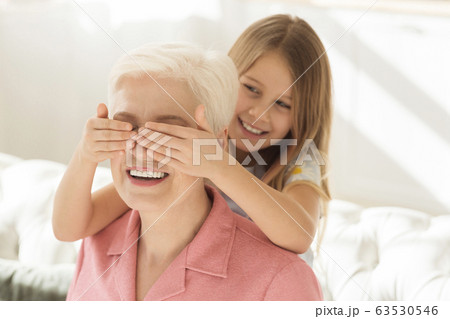 Happy little girl closing grandmother's eyes from behind, surprising her at home 63530546