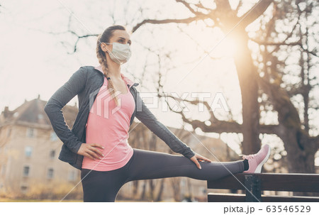 Woman wearing face mask stretching on a bench outdoors Woman wearing face mask stretching on a bench outdoors 63546529