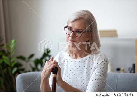 Pensive older lady in eyeglasses sitting on couch with cane. Pensive older lady in eyeglasses sitting on couch with cane. 63548848