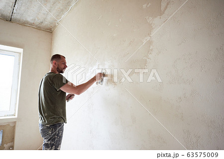 Guy is making repairing in apartment by putting putty on the walls. 63575009