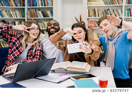 Selfie time in library. Four cheerful, smart and successful international students with smiles and crazy funny hand and face gestures, making grimace, are posing for selfie shot. 63579747
