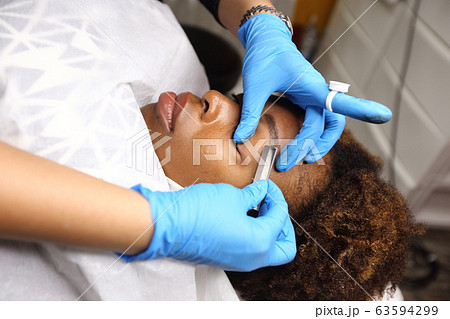 Professional Beautician woman applying Tottoo, Brow Microblading to Black african customer eyebrows to make permanent color makeup or tattooing line look real in beauty salon, copy space for text log Professional Beautician woman applying Tottoo, Brow Microblading to Black african customer eyebrows to make permanent color makeup or tattooing line look real in beauty salon, copy space for text log 63594299