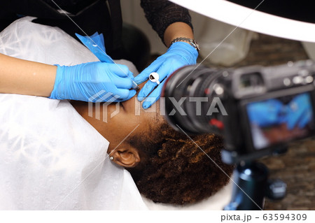 Professional Beautician woman applying Tottoo, Brow Microblading to Black african customer eyebrows to make permanent color makeup or tattooing line look real in beauty salon, copy space for text log 63594309