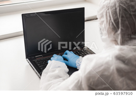 Girl in medical mask and white transparent protective suit sits in quarantine at home and works at computer at the table. Designer, artist, architect at remote work in a pandemic covid. 63597910